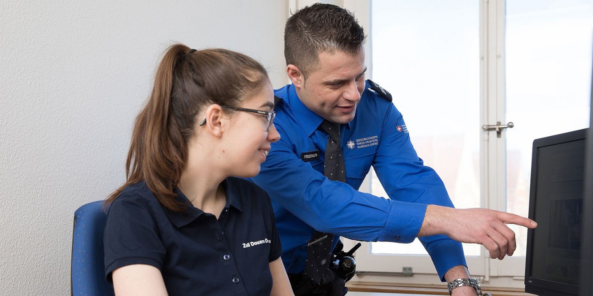 Zwei Personen, die für das BAZG arbeiten der Mann im blauen Hemd zeigt der Frau im schwarzen T-Shirt etwas auf dem Computer